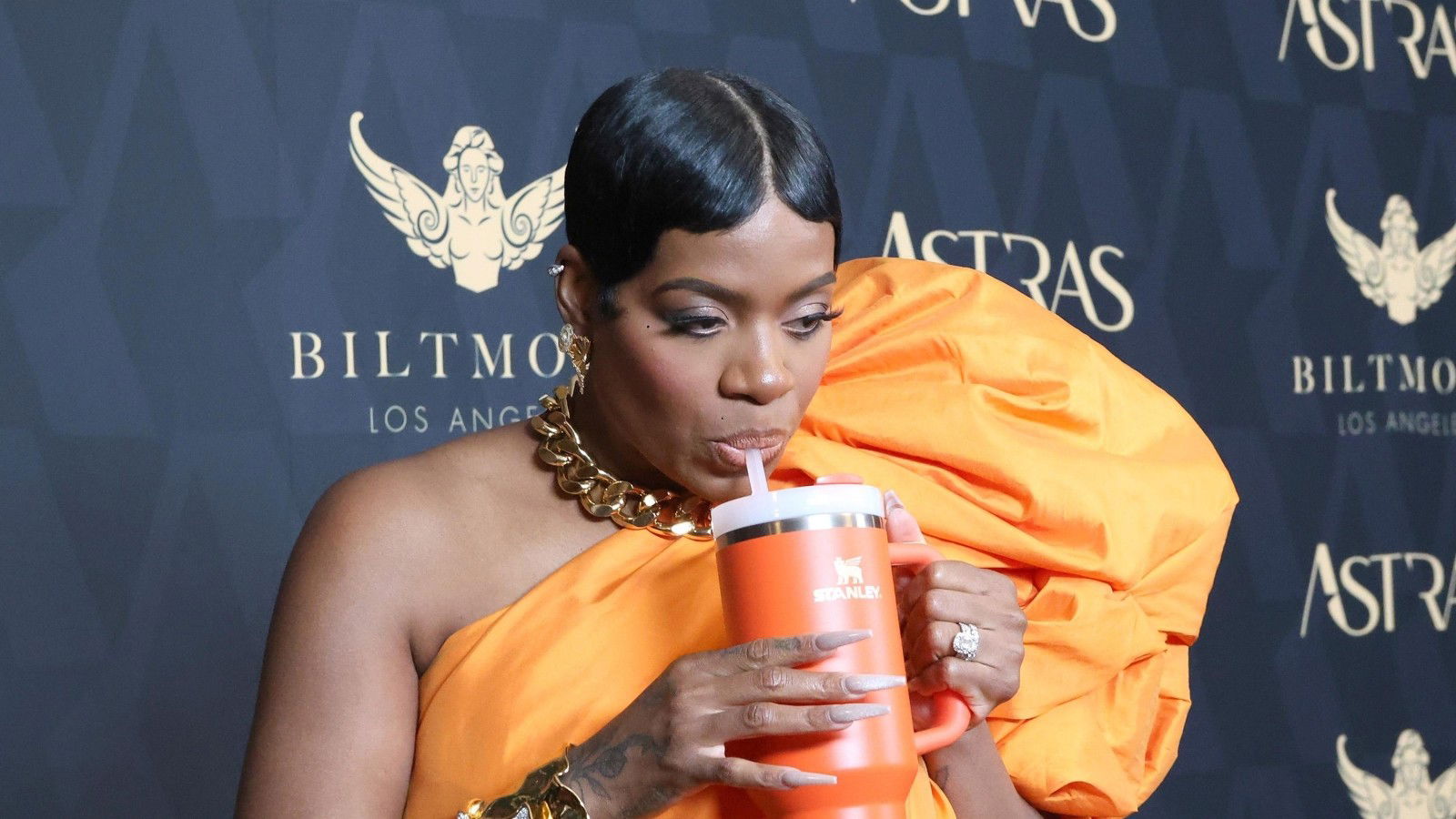 Das Bild zeigt eine Frau auf einem roten Teppich-Event. Sie trägt ein auffälliges, orangefarbenes Kleid mit voluminösem Ärmel und kombiniert es mit einer goldenen Statement-Kette. In der Hand hält sie einen orangefarbenen Stanley Cup mit Strohhalm, aus dem sie gerade trinkt. Der Kontrast zwischen glamourösem Outfit und praktischem Becher sorgt für einen originellen, modischen Auftritt.